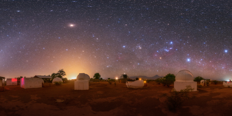 A group of observatories in-front of a dark sky. An arc of light curves from the bottom left. Several stars are visible.