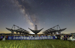 A group of people stand in-front of radio telescopes with the Milky Way in the sky behind