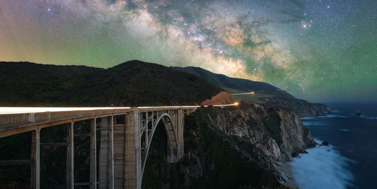The Milky Way looms over a bridge on a steep coastline. To the upper right is the bright, deep red star Antares