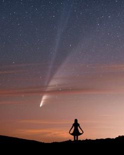 A woman in silhouette appears to greet a comet that appears behind bands of light cloud