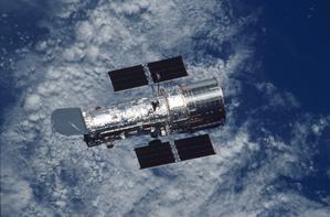 The silver-coloured Hubble Space Telescope with the blue ocean and white clouds of the Earth visible underneath.