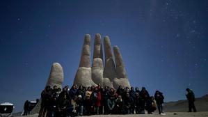 A group of people stand in-front of a giant hand scuplture before a starry sky.