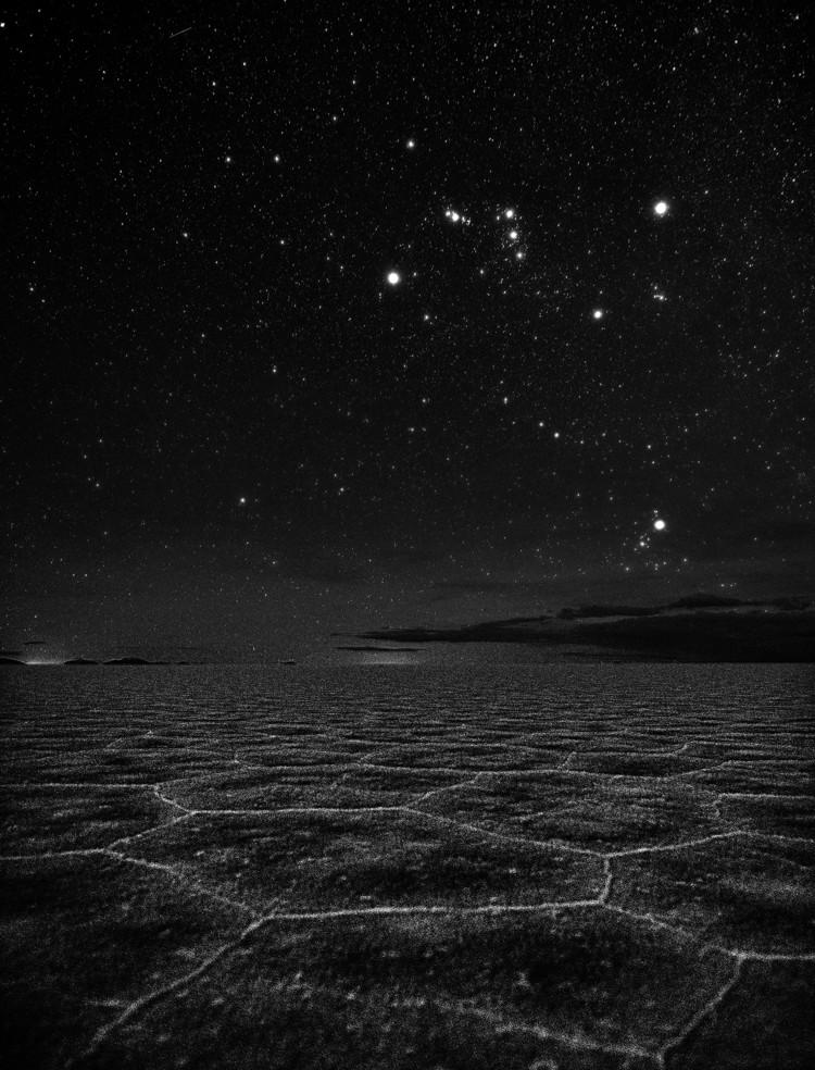 Above a flat, cracked landscape, Orion is shaped like a bow tie. Just above the horizon is a diffuse cluster of bright stars.