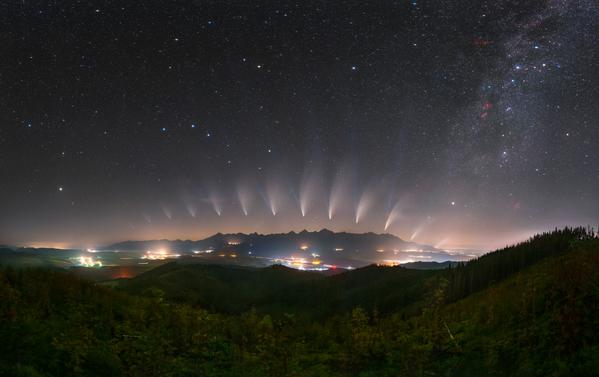 Multiple image of a comet, its tail pointing away from the horizon, form an arc in the night sky over an urban area