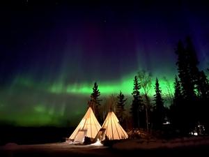 Two illuminated teepees in a subarctic forest. Green bands of aurorae light up the sky.