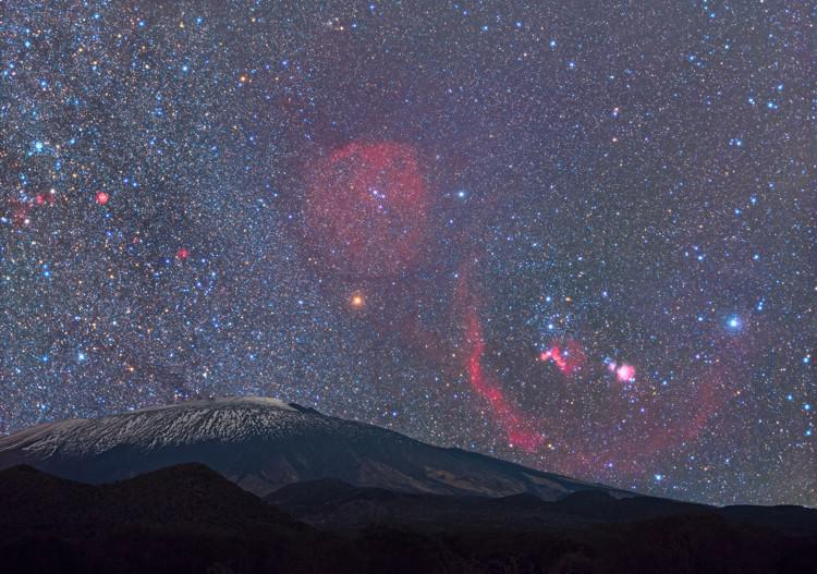Above a volcano, a bow-tie-shaped Orion is peppered with bright sweeps of nebular gas