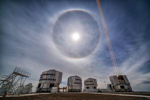 Le quattro cupole del telescopio VLT. Una cupola è aperta e spara raggi laser. La luna in cielo ha un anello di luce intorno.