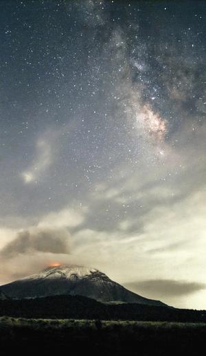 The Milky Way behind patchy clouds. Below a red glow comes from the summit of a snowy volcano.