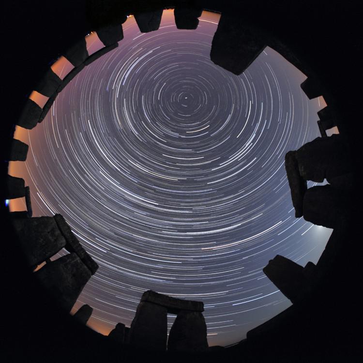 An all-sky image. Above a ring of stones shaped like door-frames, the bright curved paths of star trails seem to form circles