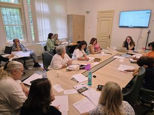 Several people sit round a table taking notes as a presenter at the head of the table shows a presentation on a screen.