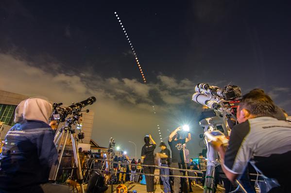 Two astronomers view a lunar eclipse. Between them in the partly cloudy sky, a series of bright circles trace a curved path