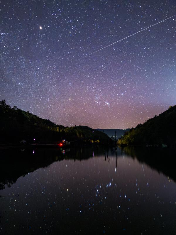 A starry sky is reflected in a lake in a valley. The streak of a meteor goes from the top right to the middle of the image