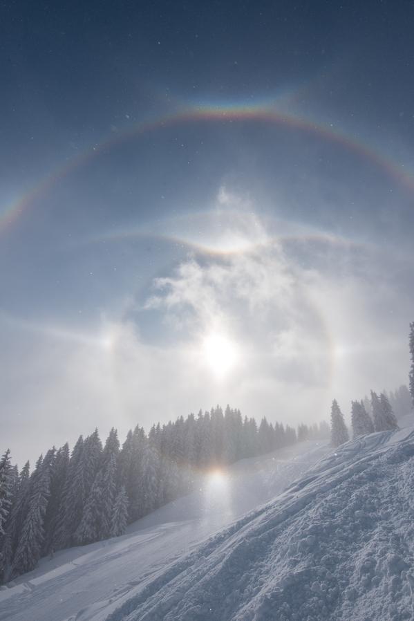 The Sun, surrounded by several bright circles and arcs, over a snowy, tree-lined landscape.